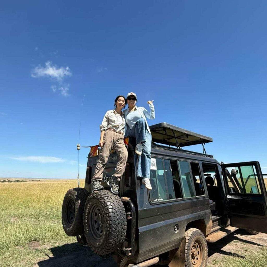 Two individuals seated atop a safari vehicle, surrounded by a vast field under a clear sky.